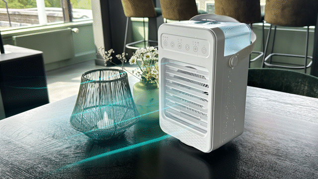 A white portable cooler fan with control buttons on top, placed on a round table next to decorative flowers and a candle holder, with green plants in the background.